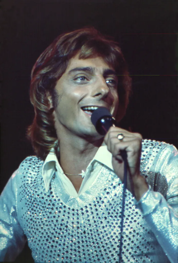 Barry Manilow singing into a microphone, wearing a shiny shirt with a star necklace.