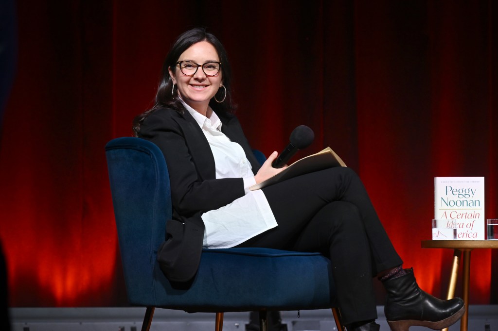 Bari Weiss speaking on stage, seated in a blue chair, holding a microphone and a book.