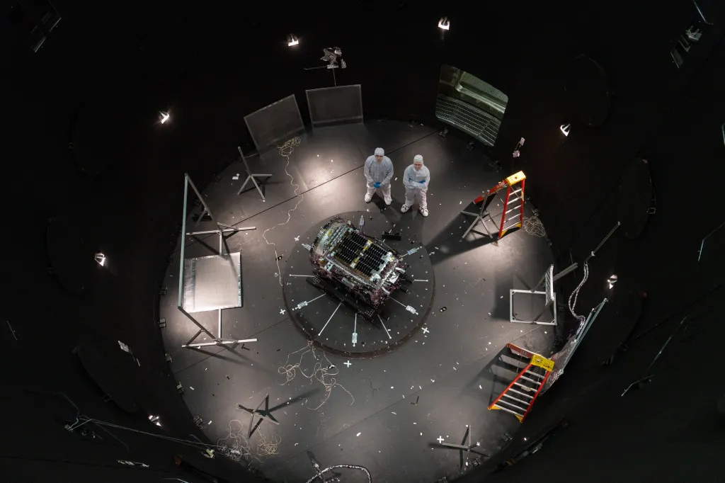 Two men in cleanroom suits standing next to a satellite in a dark test chamber.