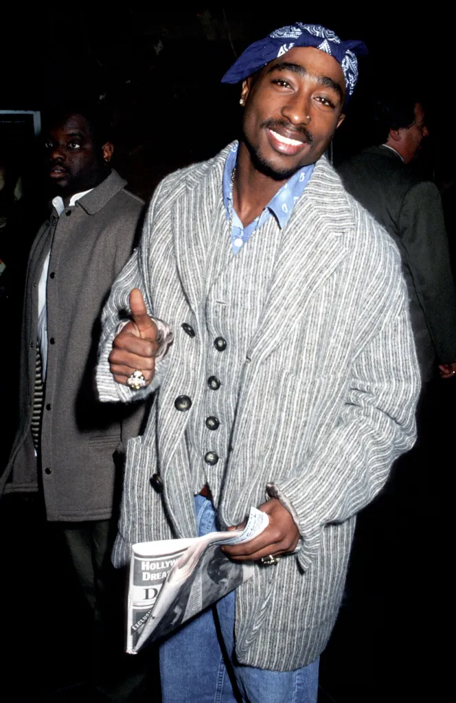 Tupac Shakur smiling and giving a thumbs up, wearing a blue bandana and a striped jacket, holding a newspaper.