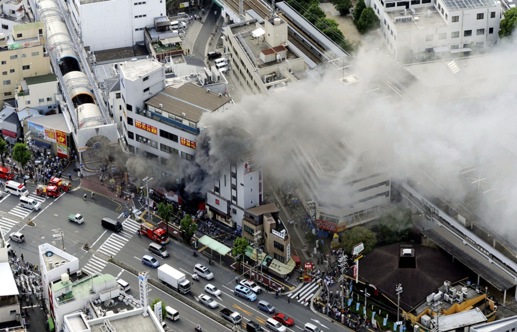 Aerial view of a deadly fire at a pachinko parlor in Osaka, Japan, with thick smoke billowing from the building, surrounded by emergency vehicles and onlookers.