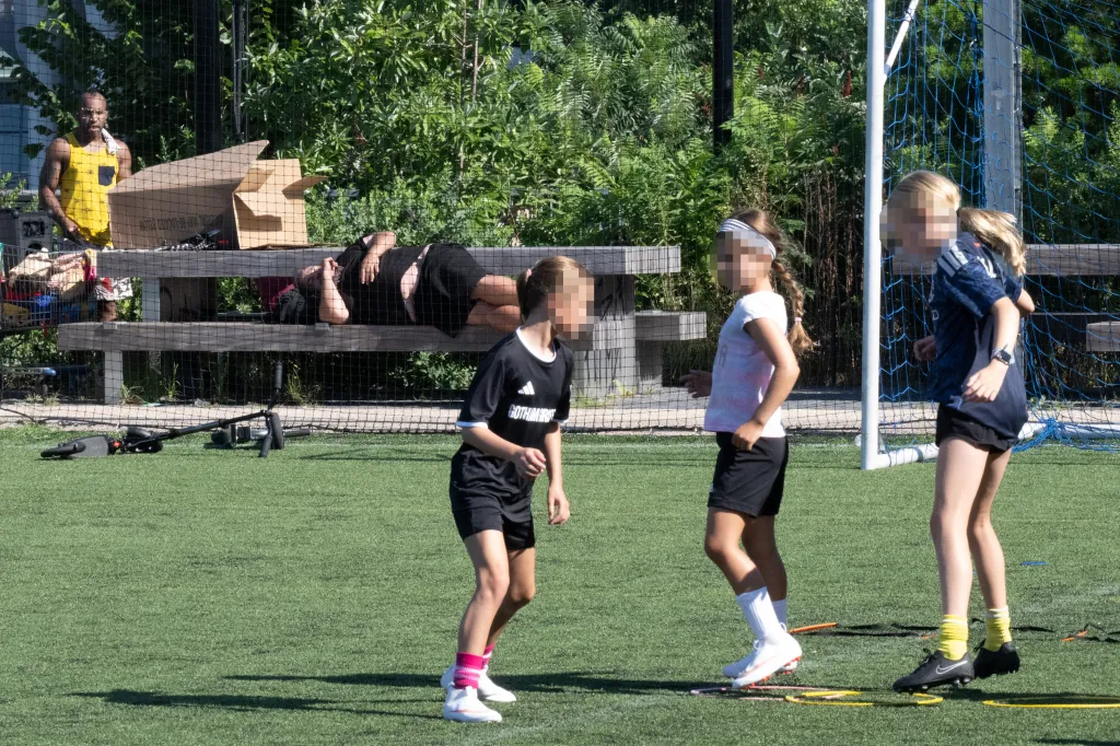 A man in a yellow tank top standing next to boxes on a bench, a person lying on the bench, and three girls on a soccer field in front of a goal.