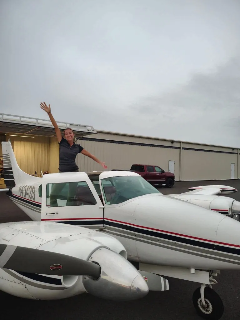 Pilot Faith Tenkley standing in the open doorway of a small white airplane with her arms raised in the air.
