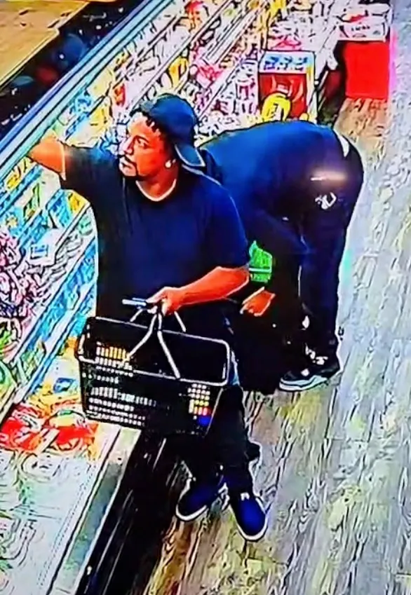 Two men shoplifting meat from a store, one holds a basket while the other stuffs a backpack.
