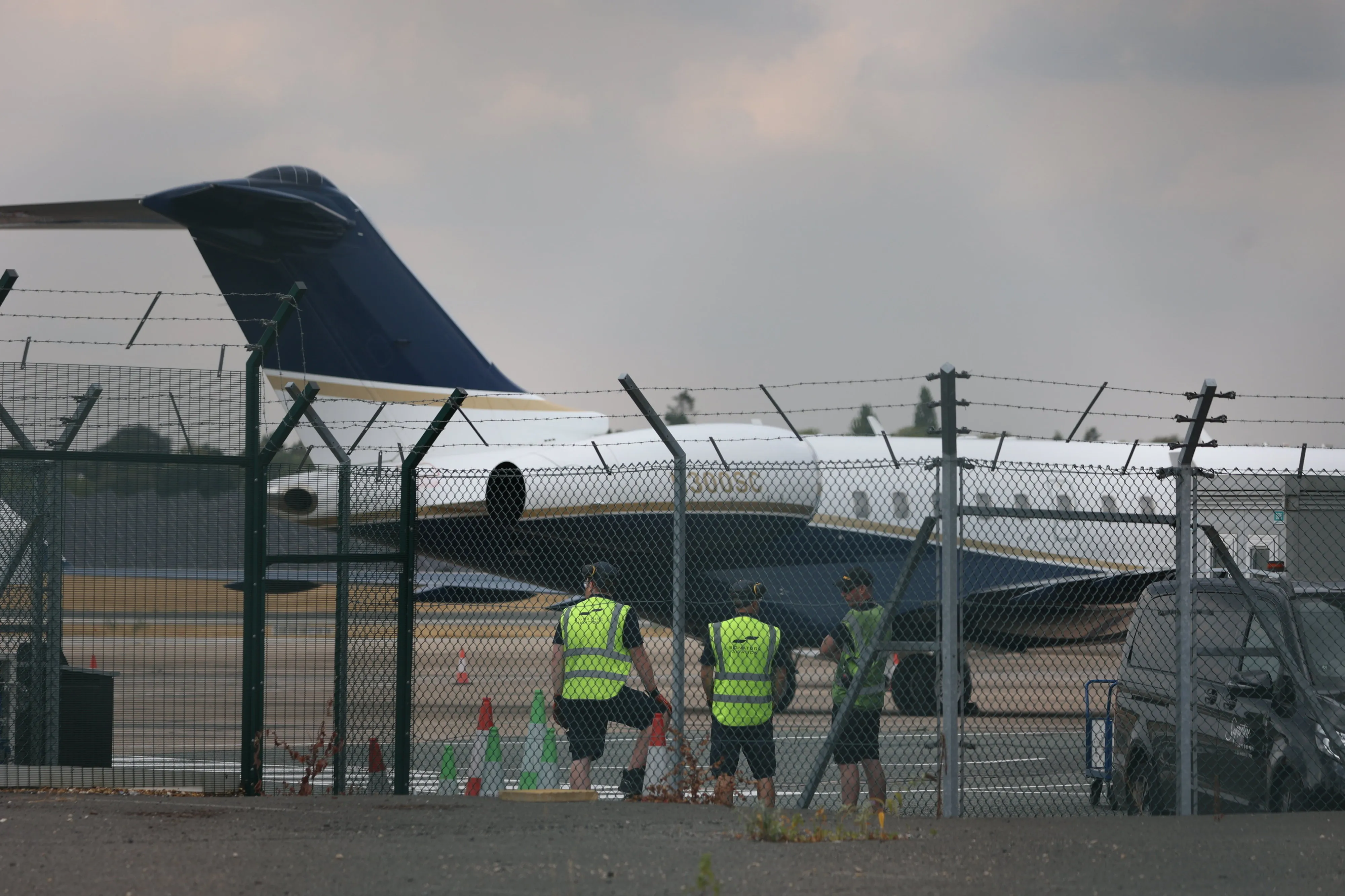 Workers stand by a fence at Birmingham Airport, with a private jet believed to have transported Prince Harry and Meghan visible in the background.