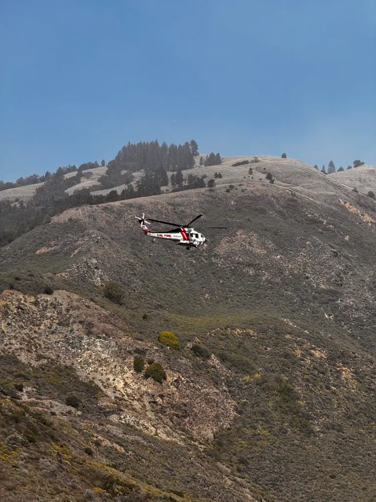 A CAL FIRE helicopter flies over a dry, brush-covered mountain in Big Sur.