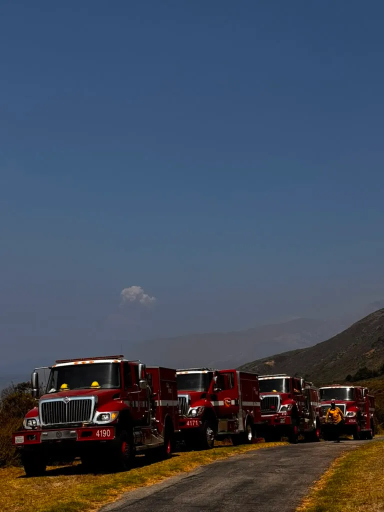 Red fire trucks on a winding road with smoke from the Plaskett Fire in the distance.