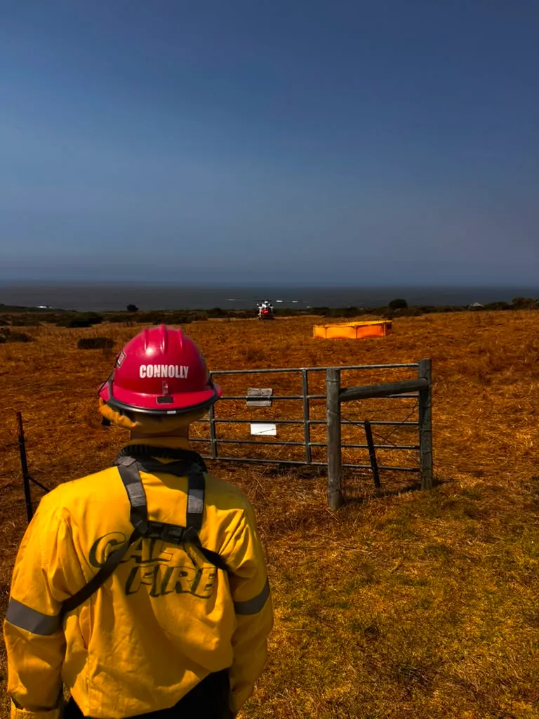 A Cal Fire firefighter wearing a red helmet looks out over dry fields towards the ocean.