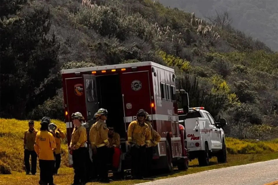 Firefighters in uniform next to a red fire truck and a white Cal Fire truck, with a brushy hillside in the background.