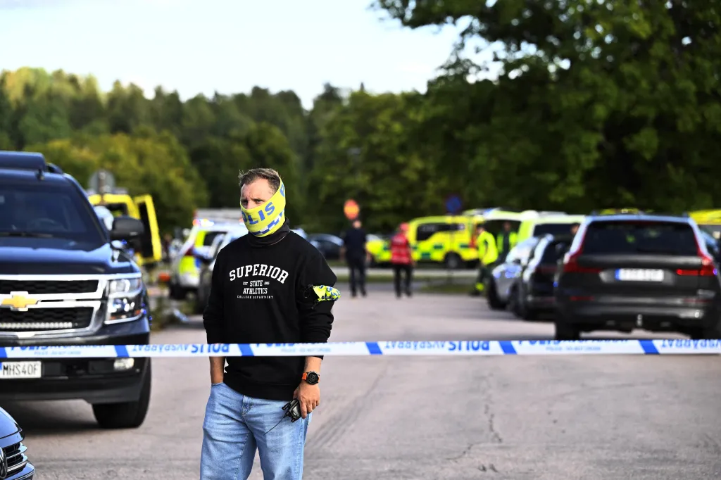 A man in a black sweatshirt and light blue jeans stands behind blue and white police tape at a high school where a sword attack occurred, with emergency vehicles and personnel in the background.