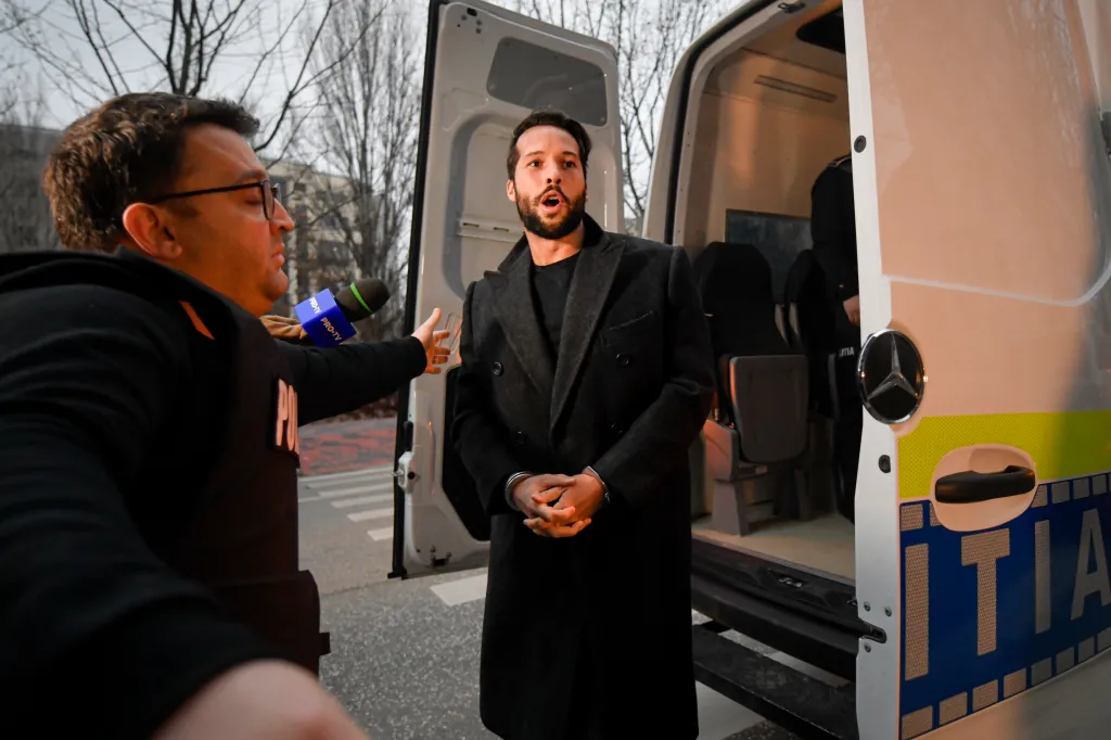Tristan Tate, handcuffed, stands in front of an open police van as a reporter extends a microphone toward him.