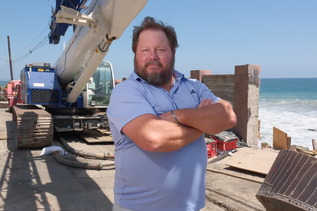 A bearded man in a blue polo shirt standing with his arms crossed in front of construction equipment and the ocean.