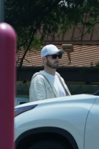 Brian Hickerson in a white hat and sunglasses, walking beside a white car.