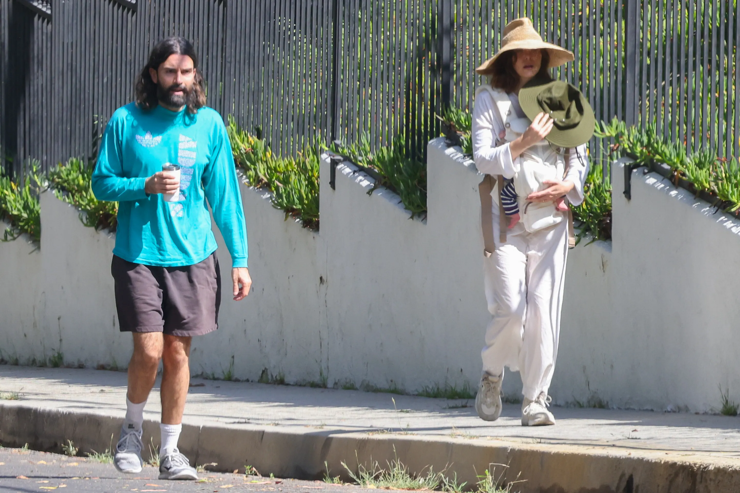 Elizabeth Olsen wearing a large straw hat while carrying a newborn in a baby carrier, and Robbie Arnett walking beside her.