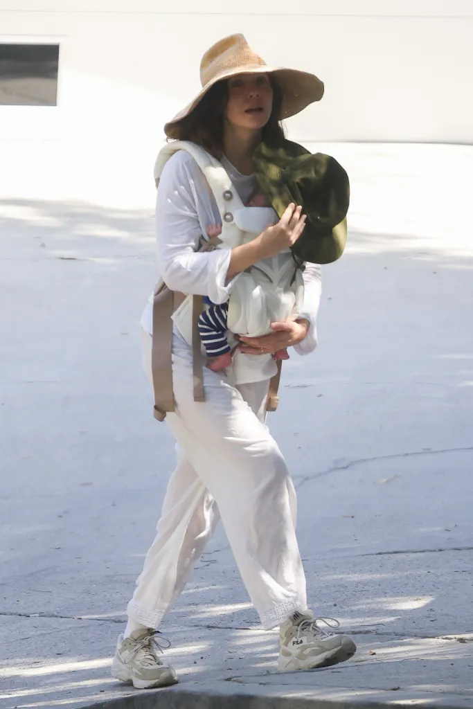 Elizabeth Olsen in a straw hat and sneakers, carrying a newborn in a baby carrier.