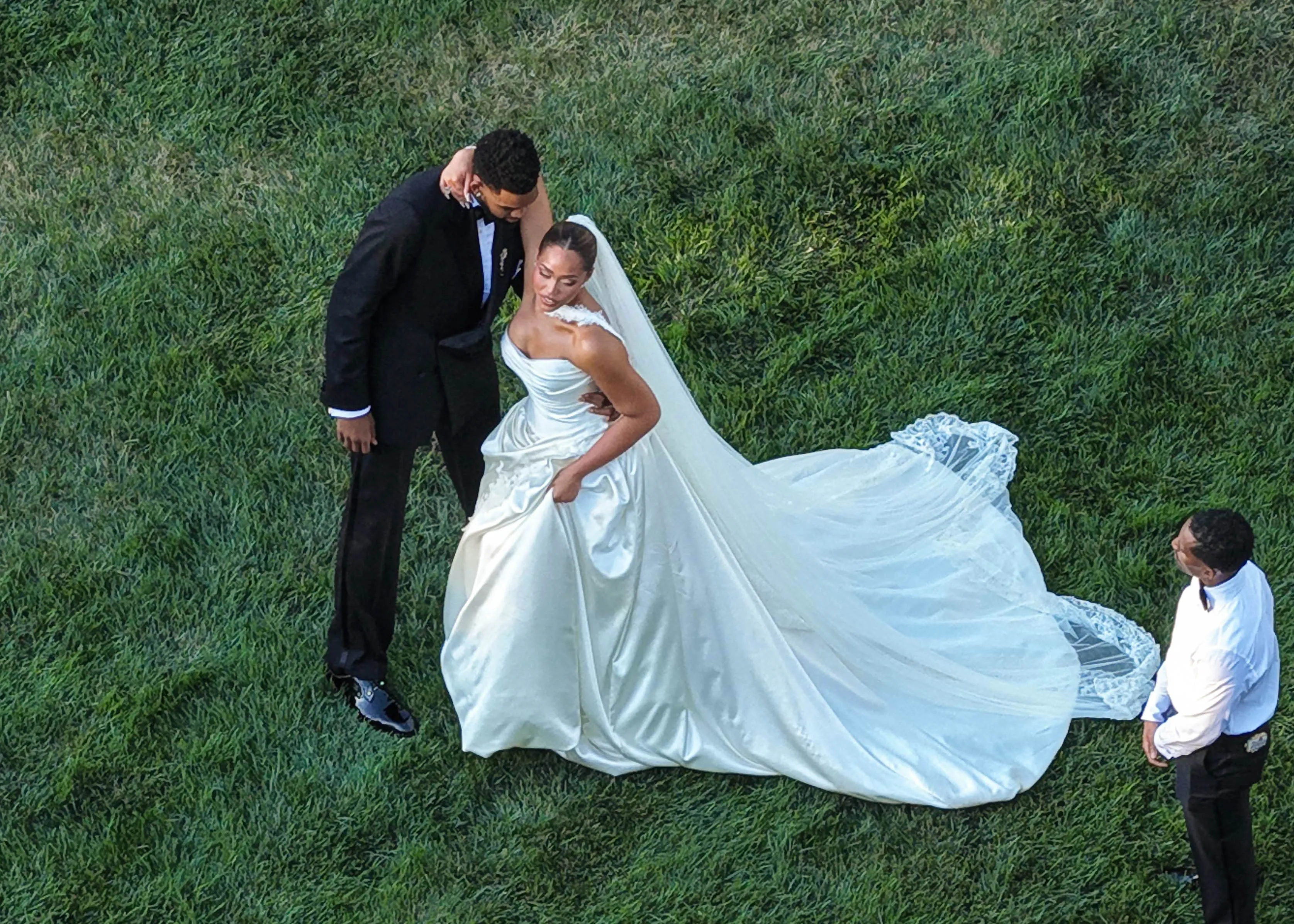 Jordyn Woods and Karl-Anthony Towns posing for wedding photos on grass, with Woods in a white dress and veil, and Towns in a black suit.