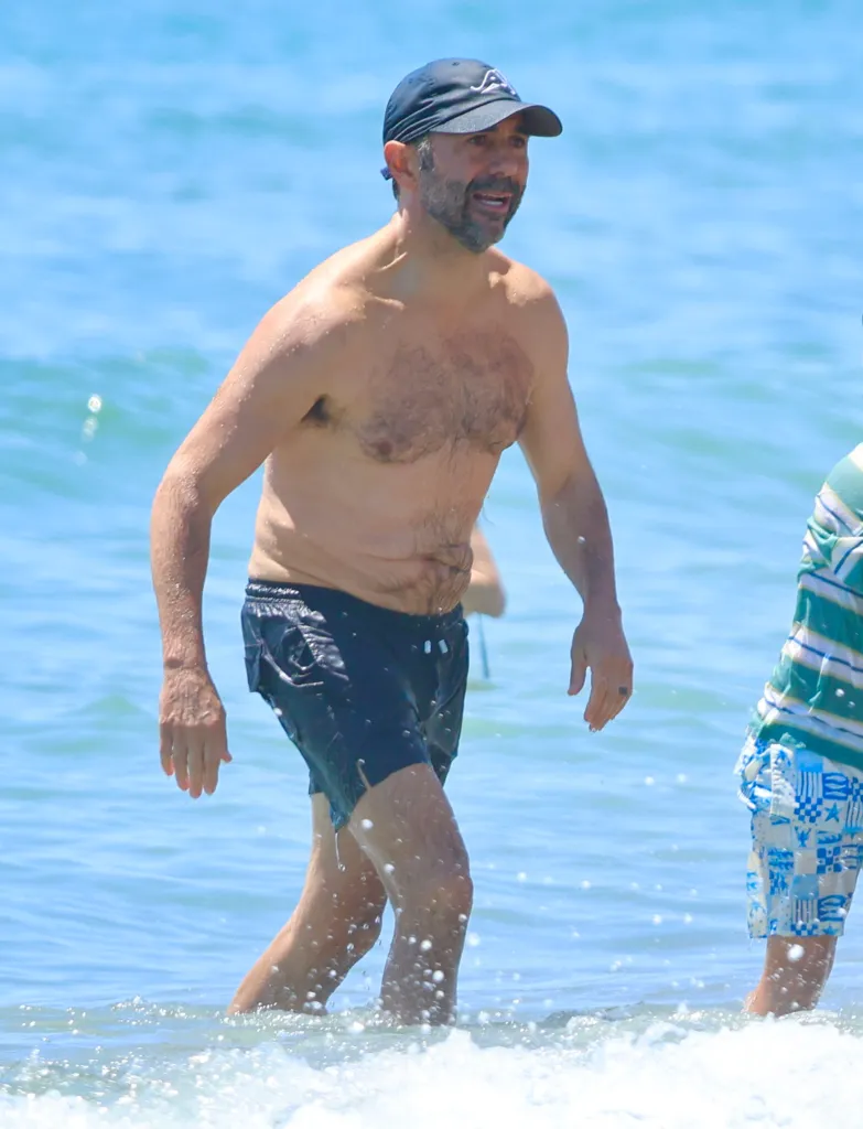 Jose Baston in dark swim shorts and a navy baseball cap walks through the shallow ocean water.