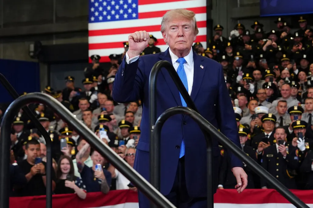 Donald Trump raising his fist to a crowd of uniformed officers and civilians with an American flag in the background.