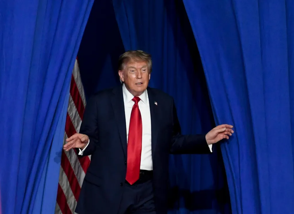 President Donald Trump emerges from behind blue curtains onto a stage, with a US flag visible.