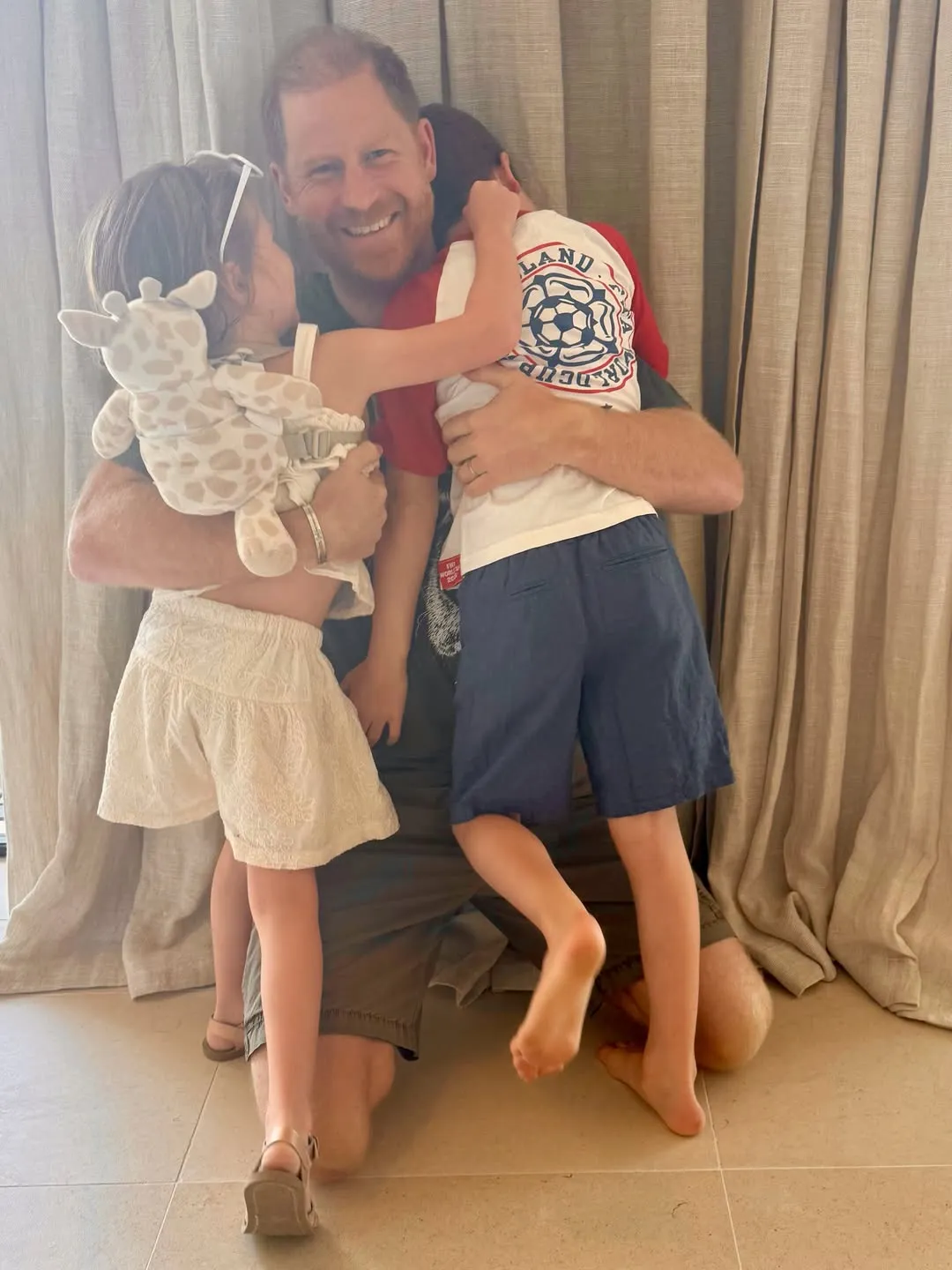 Prince Harry smiling, kneeling, and hugging his two children.