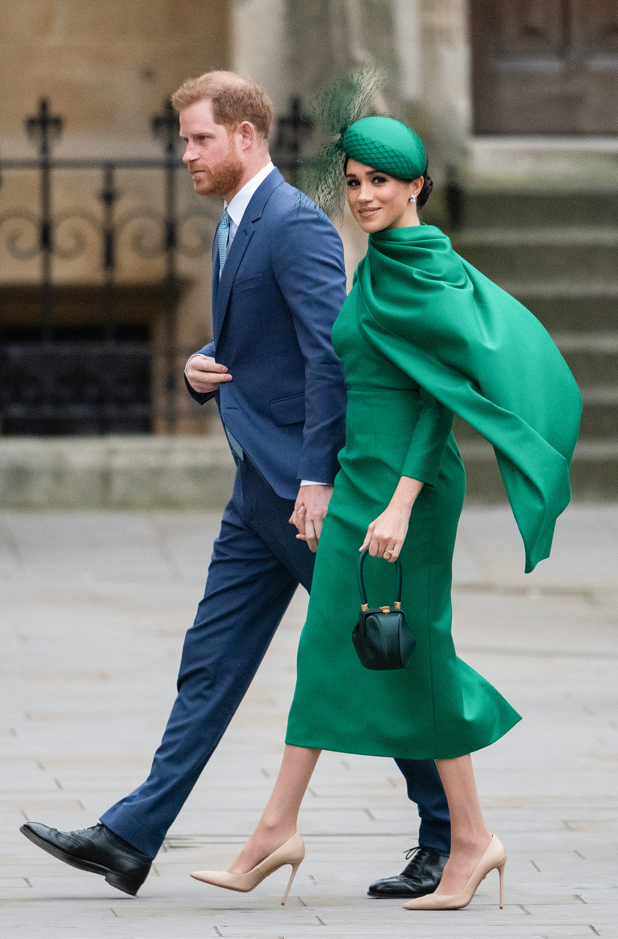 Prince Harry in a blue suit and Meghan Markle in a green dress and matching hat walk together.