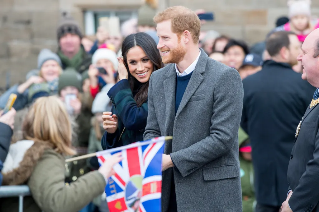 Prince Harry and Meghan Markle interacting with crowds, with Meghan smiling and adjusting her hair, and Harry reaching towards a child holding a British flag.