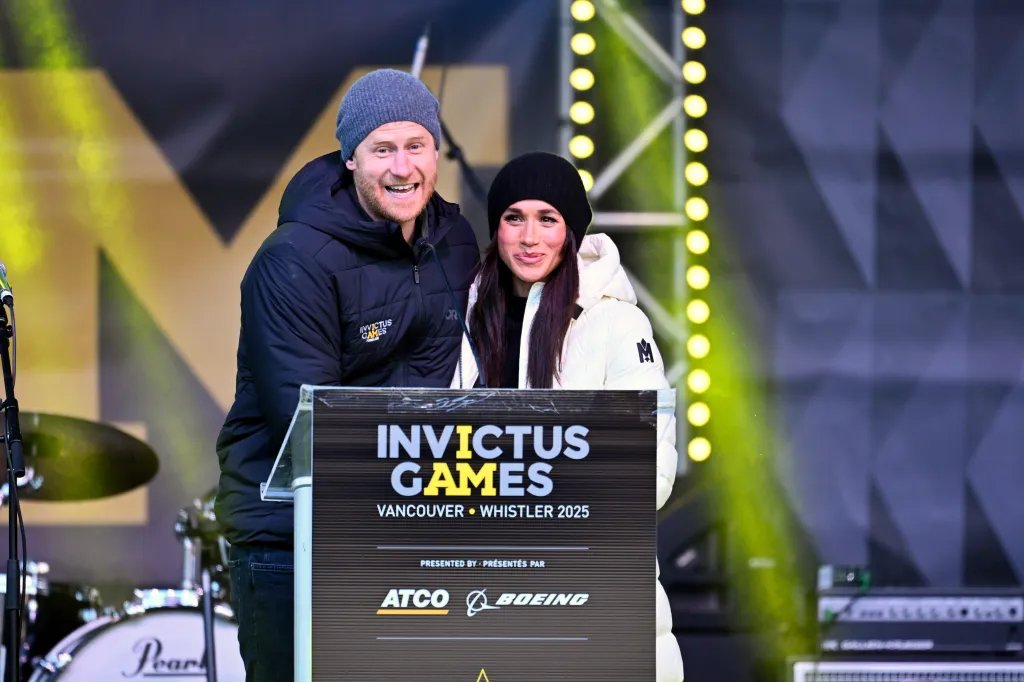 Prince Harry, in a grey beanie hat and blue Invictus jacket, and Meghan Markle, in a black beanie and white puffer coat, speaking at the 2025 Invictus Games in Vancouver and Whistler.