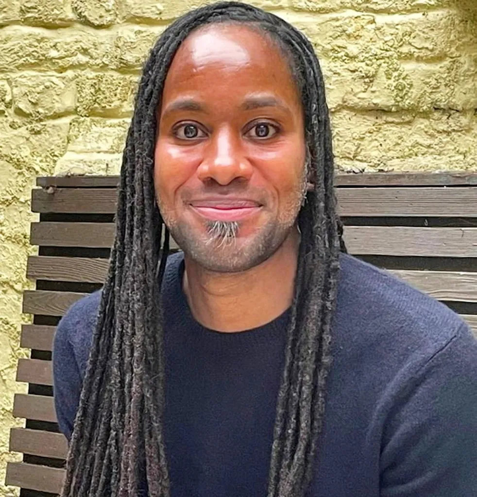 Professor Jason Arday, a Black man with dreadlocks and a goatee, smiles at the camera while wearing a navy blue sweater.