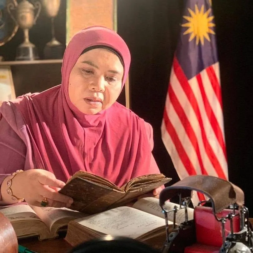 Professor Solehah Yaacob reading a book at a desk with a Malaysian flag behind her.