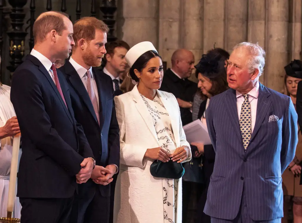 Prince Charle, in a blue suit, talking with Meghan, Duchess of Sussex, in a white coat , white hat, black and white dress, while Prince William, in a blue suit, talks with Prince Harry, in a blue suit.