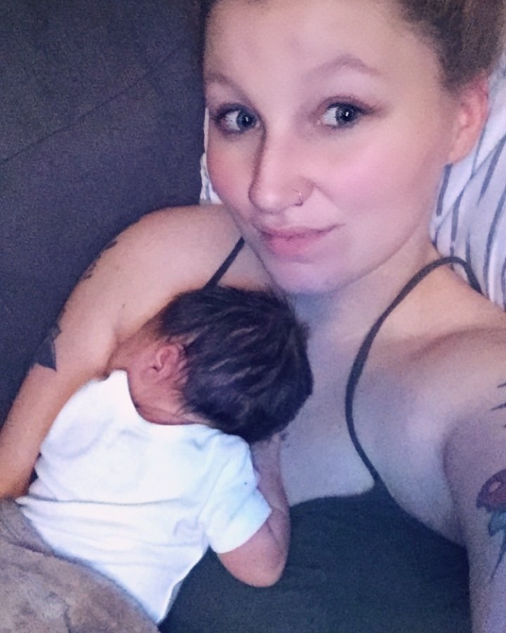 A woman with light hair and blue eyes smiles while holding a baby whose head is resting on her chest.