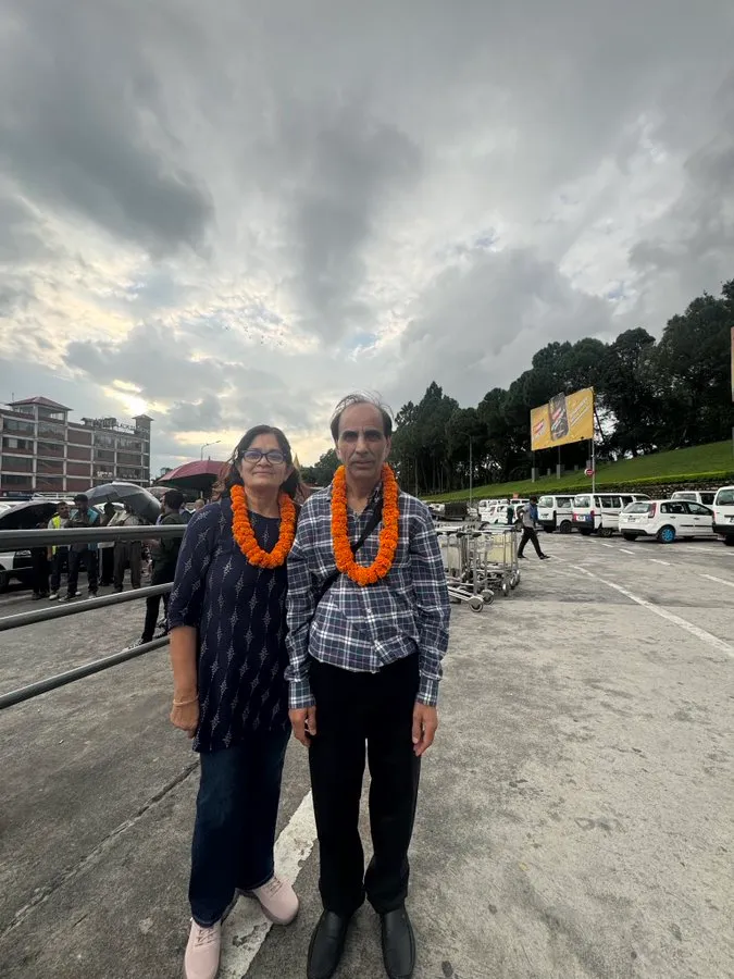 Ramesh and Neelam Sharma, wearing marigold garlands, standing outdoors against a cloudy sky.