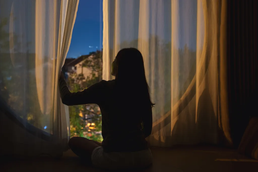 A woman looking out a window at night, holding back a sheer curtain.