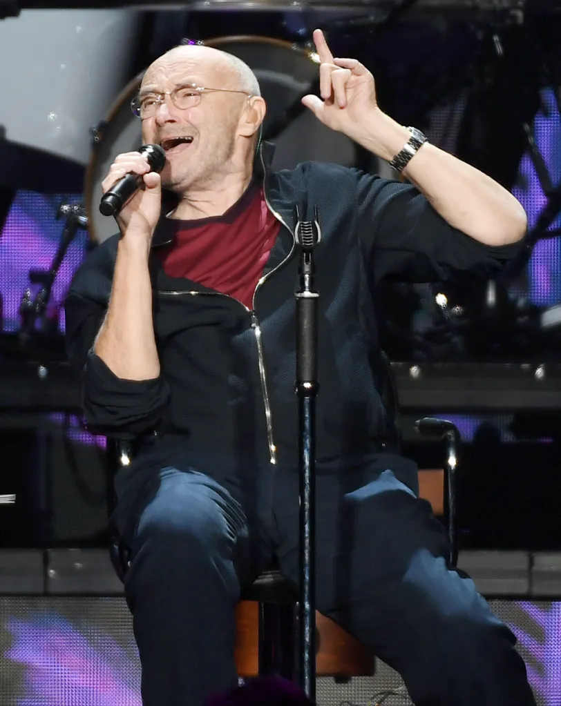 Phil Collins performing on stage, wearing glasses, a dark red shirt, and a dark blue tracksuit, holding a microphone in one hand and pointing upwards with the other.