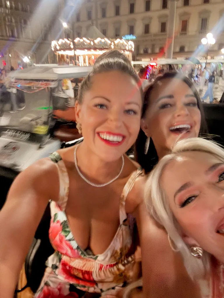 Three women smiling at the camera at night with a carousel in the background.
