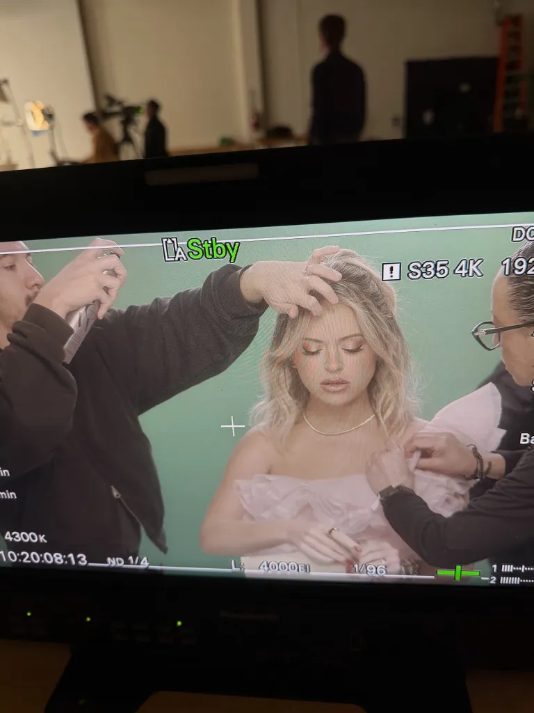 Video monitor showing a blonde woman in a pink dress being styled by two people on a green screen set.