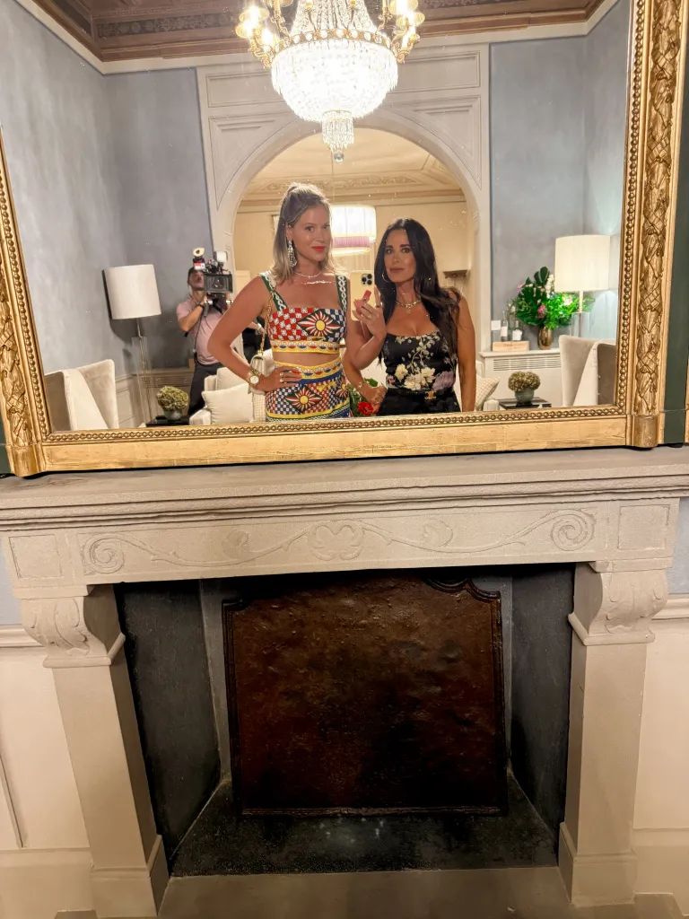 Two women reflected in a large mirror, standing above a fireplace.