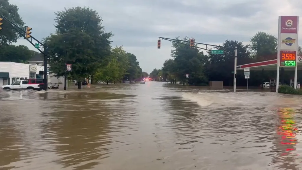 A flooded road in Cambridge City, Indiana, with cars partially submerged and a gas station on the right.