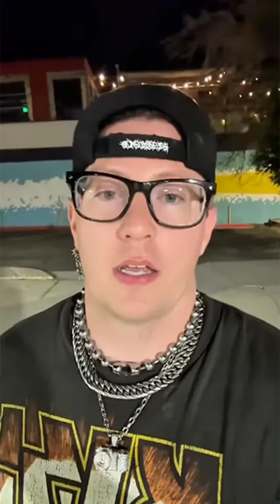 Nathan James, rock star, wearing a black cap, glasses, and layered silver necklaces, looking directly at the camera.