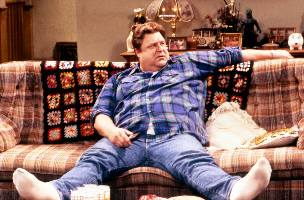 John Goodman as Dan Conner from Roseanne, sitting on a plaid sofa.
