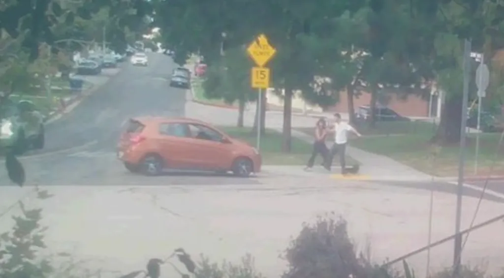 An orange car has driven onto a sidewalk and appears to be trying to hit a couple and their dog.