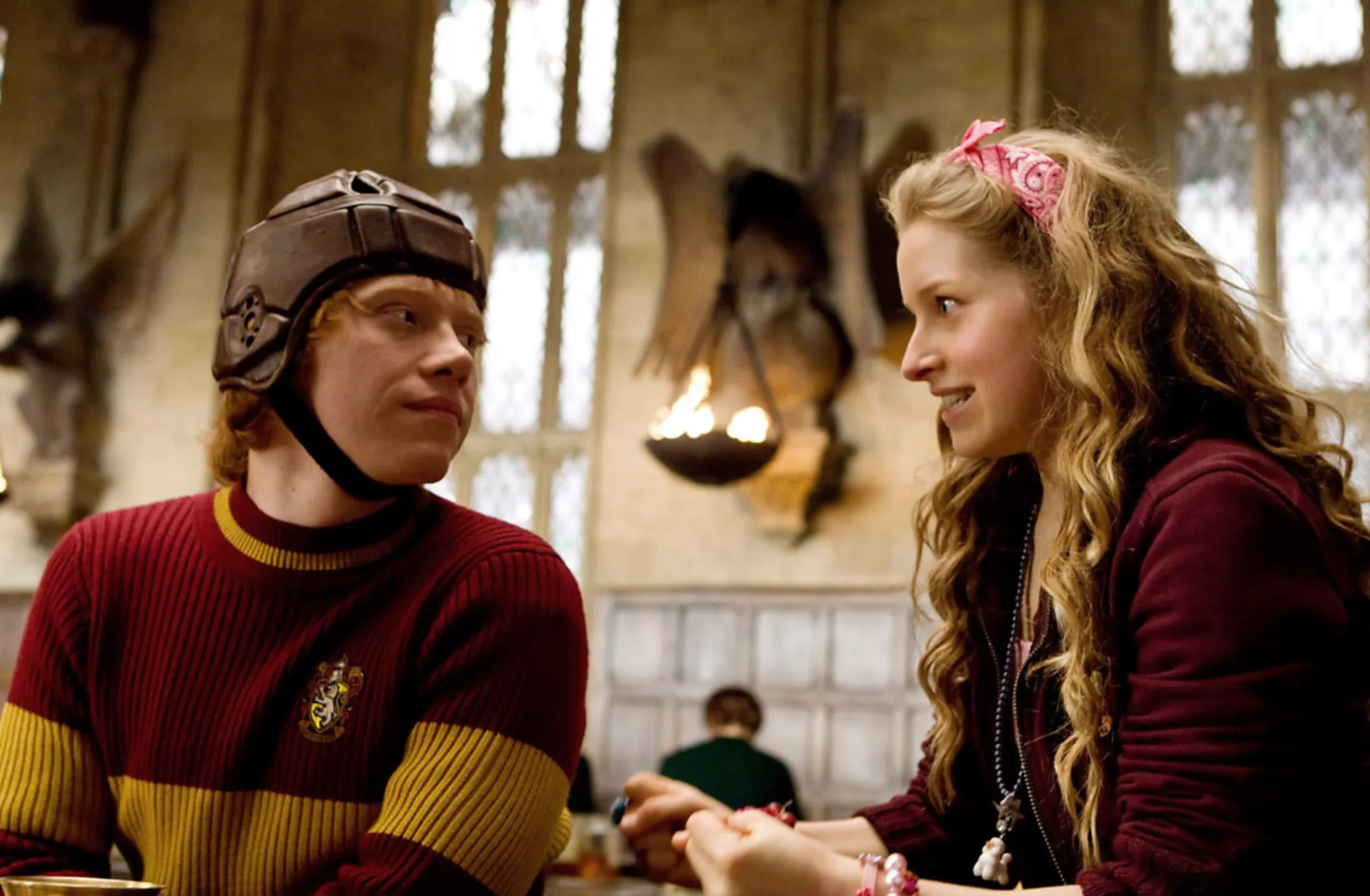 Rupert Grint as Ron Weasley in a Quidditch helmet and sweater, and Jessie Cave as Lavender Brown with wavy hair and a pink bandana, looking at each other in 