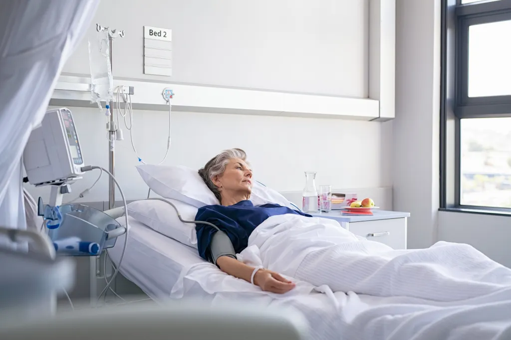A lonely elderly woman lies in a hospital bed, looking away thoughtfully.