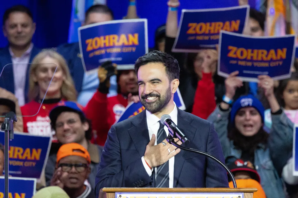 Zohran Mamdani smiling at a rally, with supporters holding