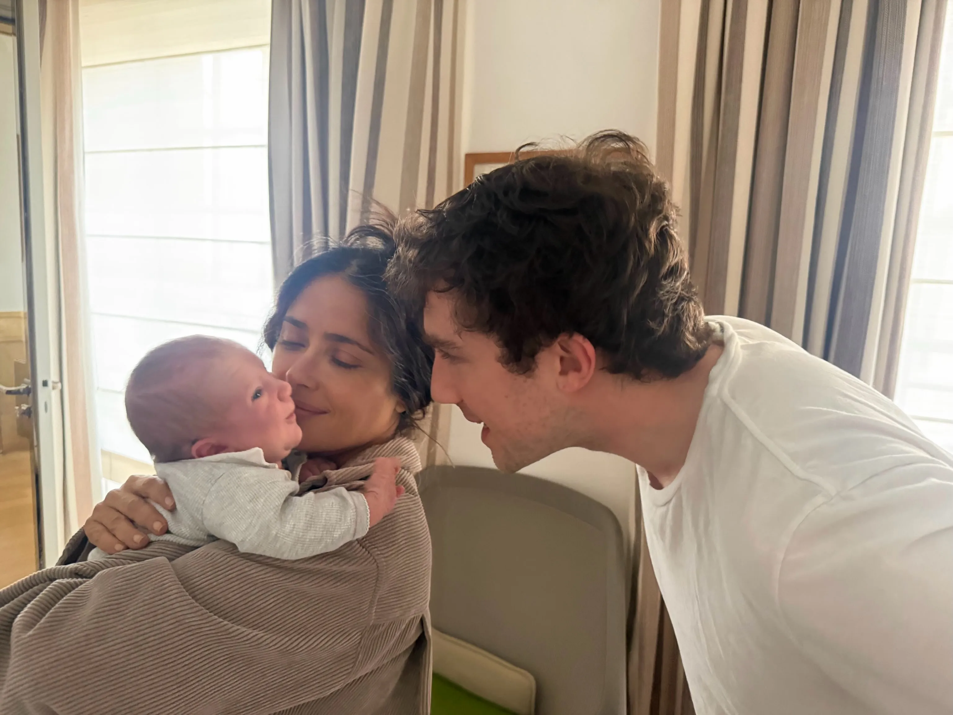 Salma Hayek holding her newborn grandson, with her stepson François Pinault Jr. smiling at the baby.