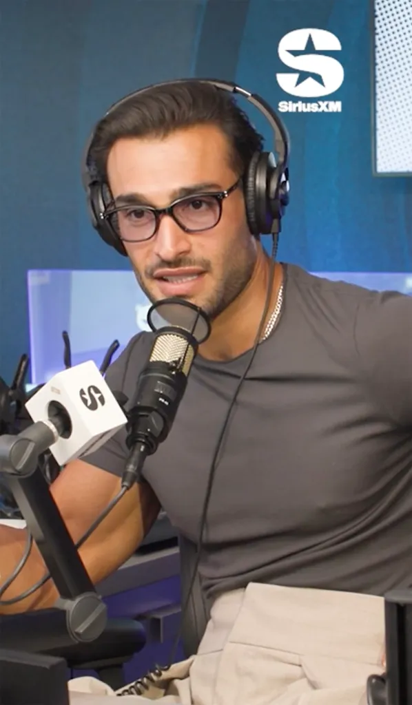 Sam Asghari wearing headphones and glasses, speaking into a microphone with a SiriusXM logo visible.