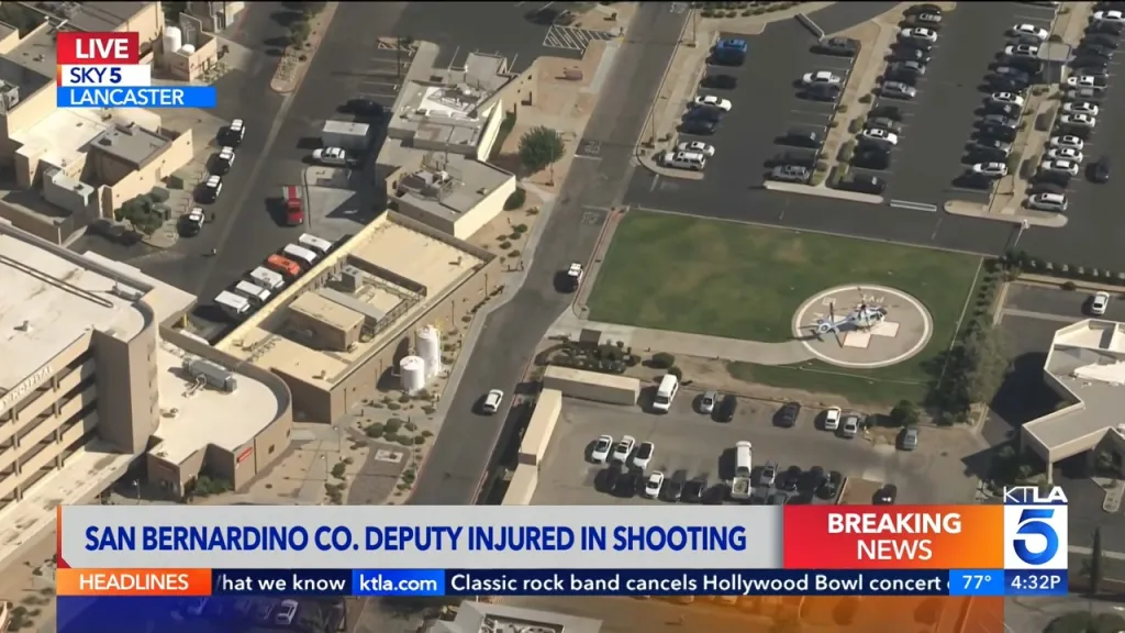 Aerial view of a hospital with a helicopter on a helipad and several emergency vehicles, with a news chyron at the bottom reading 