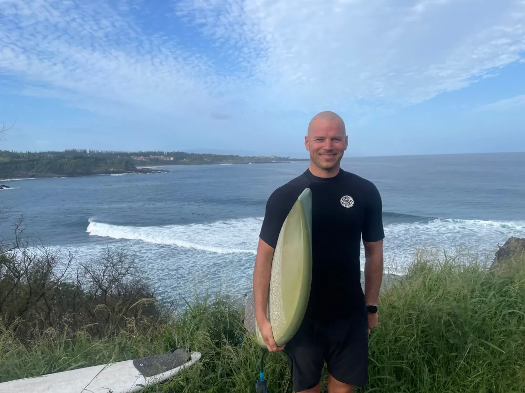 Josh Collette, a bald man in a wetsuit, holding a surfboard and smiling, with the ocean in the background.
