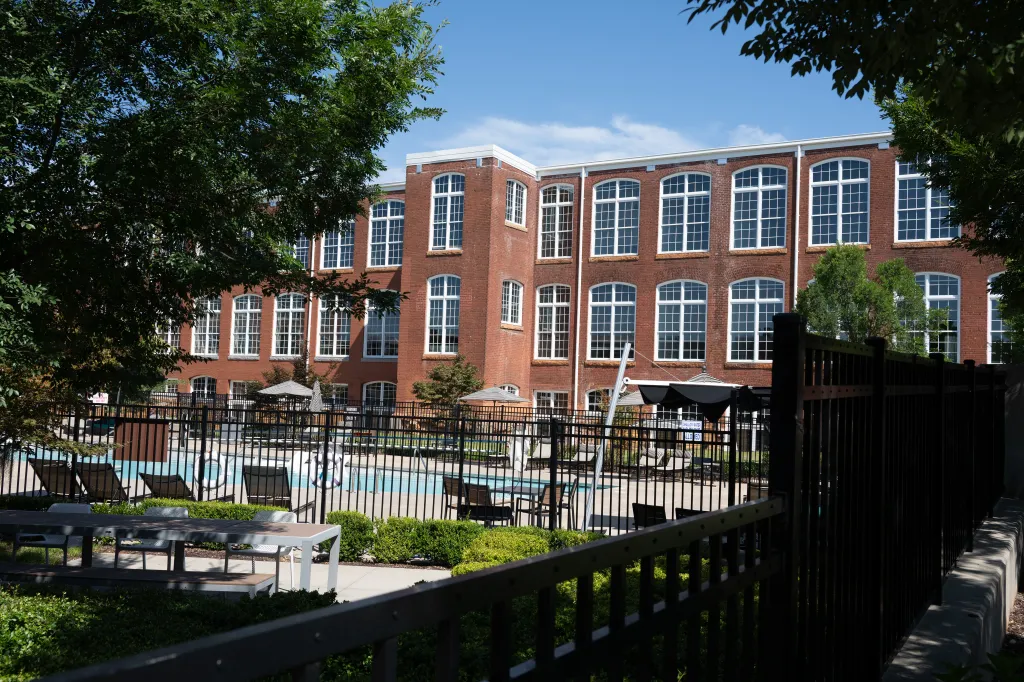 Exterior view of Judson Mill Lofts, a large brick building with many windows, and an outdoor pool area with lounge chairs and umbrellas, seen through a black metal fence.