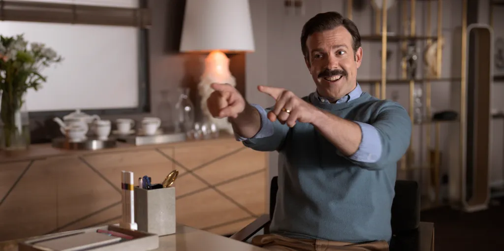 Ted Lasso (Jason Sudeikis) smiles and points at the viewer while sitting at a desk.